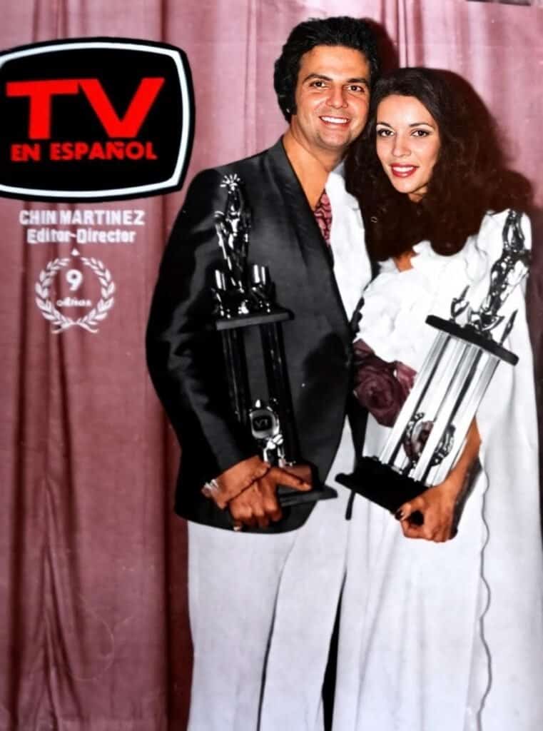 Chin Martinez holding awards at TV en Español event with a man, pink backdrop behind them.