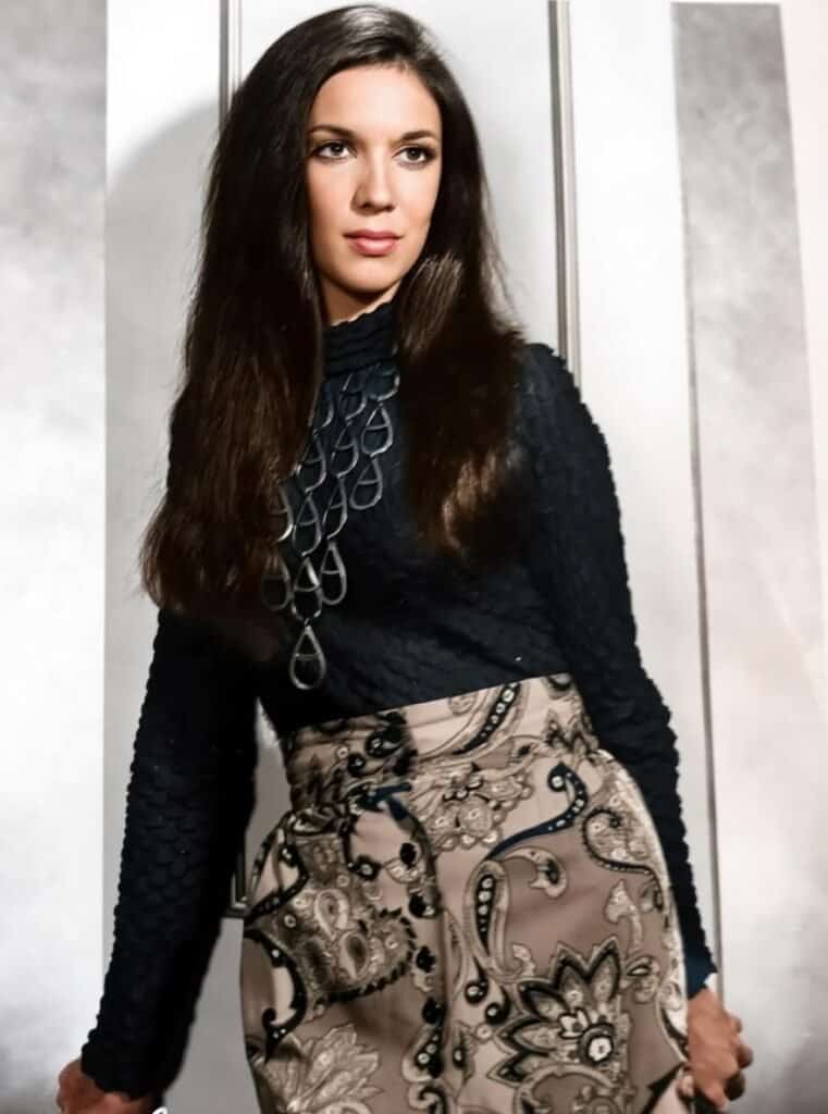 A woman with long dark hair wearing a navy blue top and patterned beige skirt, standing against a mo.