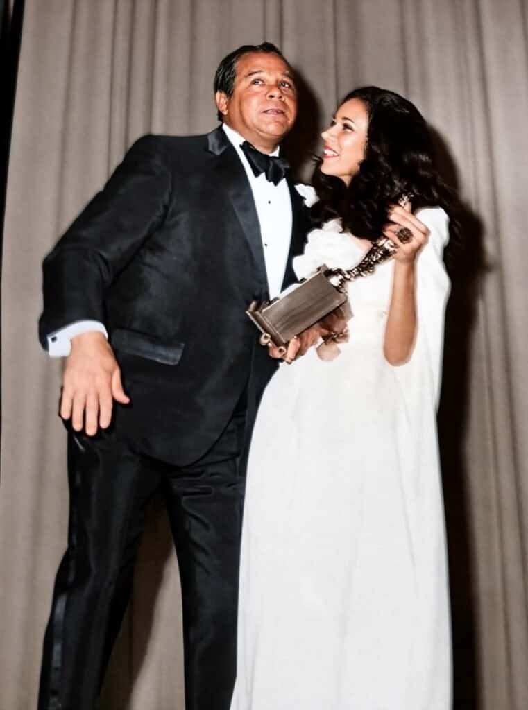 Elegant couple in tuxedo and white gown at formal event, smiling and holding an award.